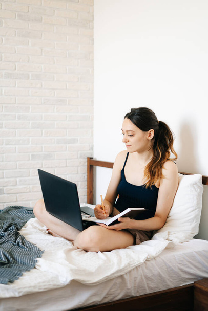 Європейка, використовуючи ноутбук, вивчаючи інформацію в Інтернеті в ліжку, пише нотатки в своєму щоденнику, в той час як вірус знаходиться в стані квартину. cozy workplace, e-learning concept - Фото, зображення