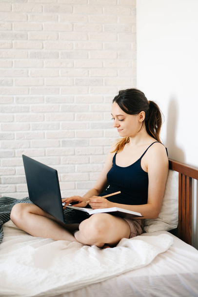 Avrupalı bir kadın, dizüstü bilgisayar kullanarak, yatağında internetten bilgi öğrenirken, virüs karantinadayken günlüğüne notlar yazıyor. Rahat bir iş yeri, e-öğrenim konsepti - Fotoğraf, Görsel