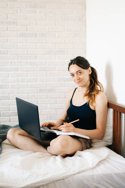 Mujer europea, el uso de un ordenador portátil, el estudio de la información en Internet en su cama, escribe notas en su diario, mientras que el virus está en cuarentena.
 - Foto, imagen