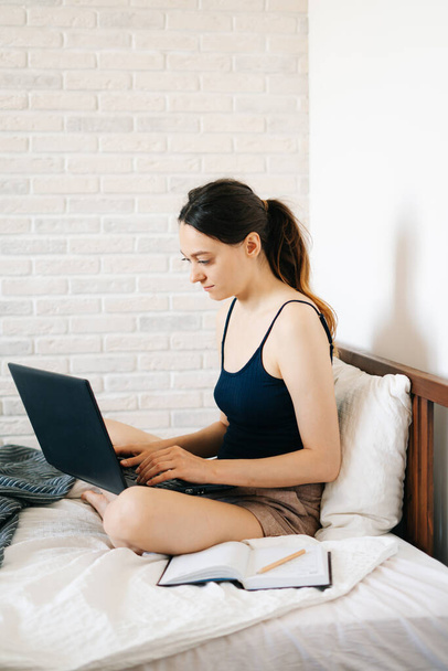 Μια νεαρή γυναίκα, χρησιμοποιώντας ένα φορητό υπολογιστή, επικοινωνεί μέσω του Διαδικτύου με έναν πελάτη στο κρεβάτι της, ενώ ο ιός είναι σε καραντίνα. Άνετος χώρος εργασίας γραφείου, Απομακρυσμένη εργασία, e-learning έννοια - Φωτογραφία, εικόνα