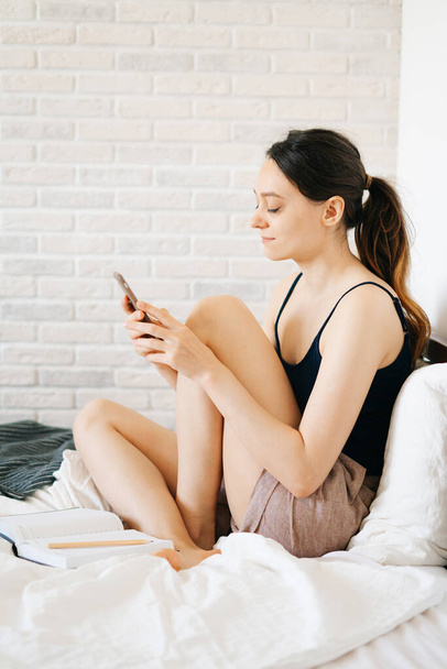 Młoda Europejka, korzystając ze smartfona, rozmawia ze znajomymi w łóżku podczas samoizolacji.Przytulne miejsce pracy, koncepcja e-learningu. - Zdjęcie, obraz
