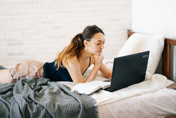 Europejka, korzystając z laptopa, studiując informacje w Internecie w łóżku, pisze notatki w pamiętniku, podczas gdy wirus jest poddany kwarantannie. Przytulne miejsce pracy, koncepcja e-learningu - Zdjęcie, obraz