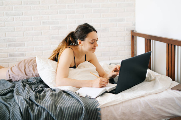 Az európai nő laptopot használ, információt tanul az interneten az ágyában, jegyzeteket ír a naplójába, miközben a vírust karanténba helyezik.Kényelmes munkahely, e-learning koncepció - Fotó, kép