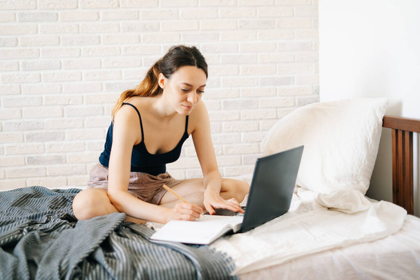 Avrupalı bir kadın, dizüstü bilgisayar kullanarak, yatağında internetten bilgi öğrenirken, virüs karantinadayken günlüğüne notlar yazıyor. Rahat bir iş yeri, e-öğrenim konsepti - Fotoğraf, Görsel