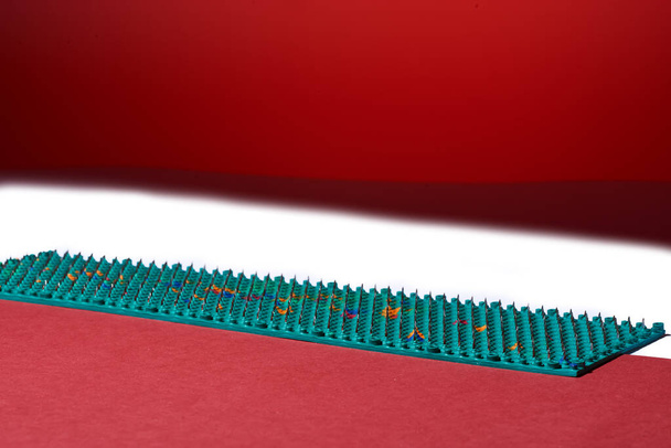 Massager in de vorm van groen tapijt met metalen spikes, naalden, ligt op rode en witte achtergronden, diagonaal. Abstractie. Voor ontspanning gezondheid. - Foto, afbeelding