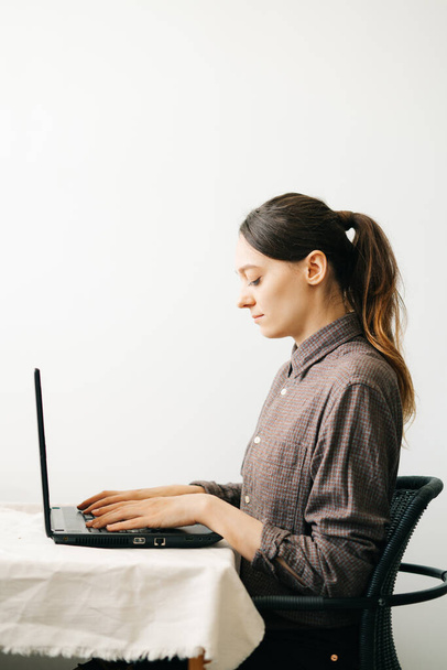 Bilgisayarında virüs karantinası sırasında evden çalışan genç bir Avrupalı kadın var. Kız minimalist bir iç mekanda beyaz bir masada oturuyor. Rahat bir iş yeri, uzaktan çalışma konsepti., - Fotoğraf, Görsel