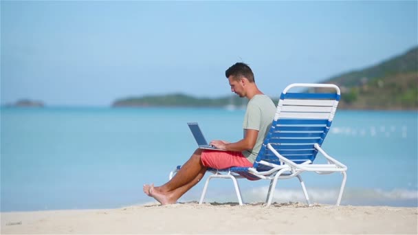 熱帯のカリブ海のビーチでノート パソコンを持つ若者。コンピューターとサンベッドの上に座って、ビーチで働く男 - 映像、動画
