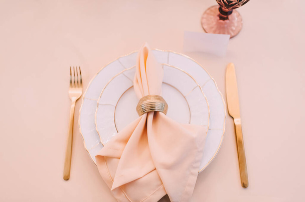 Banquet, restaurant. Table setting. White plates with a gold rim, golden cutlery, glasses. Peach-colored napkin with a golden ring.  - Zdjęcie, obraz