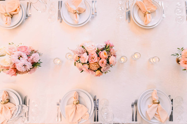 Banquet, restaurant. Table setting. White plates with a gold rim, golden cutlery, glasses. Peach-colored napkin with a golden ring. Peach pink floral arrangements - Foto, Imagen