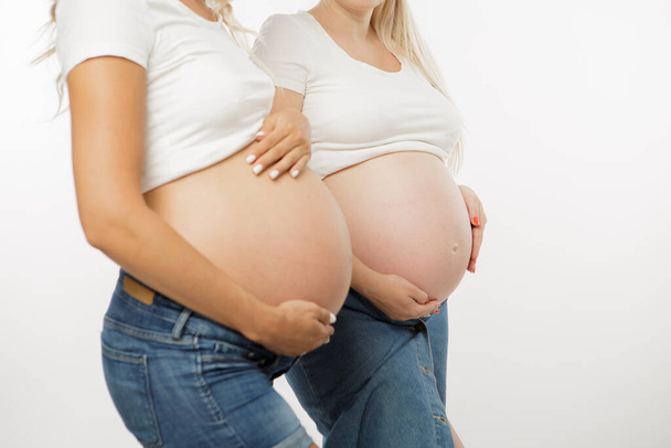 Две беременные женщины с большими животиками. Разные желудки у беременных женщин красивой формы. Желудок выглядывает из джинсов. Одежда повседневная. Белый фон. Студийный свет. Незапланированная беременность
 - Фото, изображение