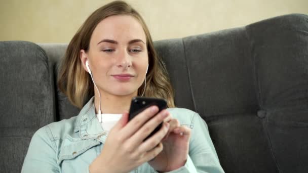 Giovane donna seduta ad ascoltare musica con gli auricolari sul suo cellulare a casa mentre si rilassa con gli occhi chiusi su un divano
. - Filmati, video