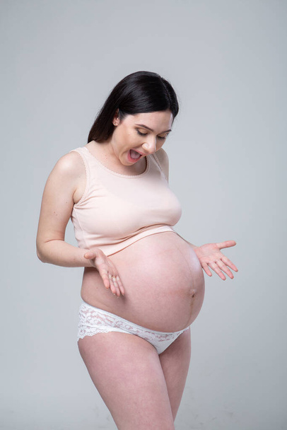Pregnant young brunette woman in lingerie. Emotional studio photo pregnant on a white background. Big round belly in a pregnant woman. Last month of pregnancy. Waiting for the baby. Beige tones.Eye contact - Φωτογραφία, εικόνα