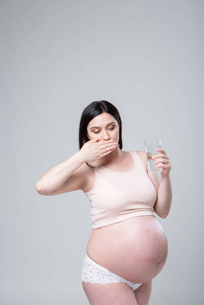 Raskaana oleva valkoihoinen nainen alusvaatteissa lasi vettä kädessä. Emotionaalinen studio valokuva raskaus valkoisella taustalla. Juoda vai olla juomatta? Viimeinen raskauskuukausi. Hän katsoo lasiin drinkin kanssa. Terveydenhuollon käsite - Valokuva, kuva