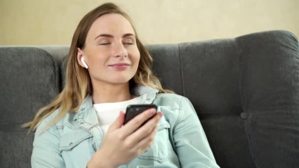 Young woman sitting listening to music with earphones on her mobile phone at home as she relaxes with closed eyes on a sofa. - Filmati, video