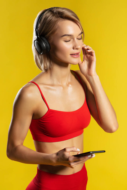 Time for music. Side view of attractive sporty blonde woman in headphones keeping eyes closed and listening music while standing against yellow background - Fotografie, Obrázek