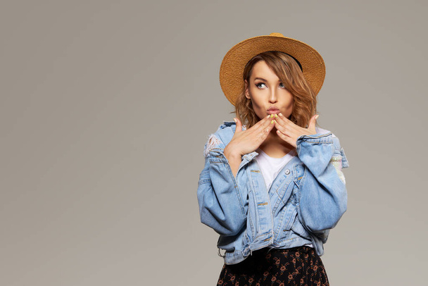 Young surprised woman model in a fashionable hat and denim jacket on a gray background. Points fingers to the sides. Space for text - Fotó, kép
