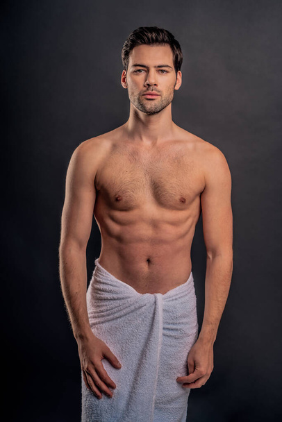 Handsome young bearded man isolated. Topless muscular man in towel is standing on grey background. Serious man looking at camera - Foto, Imagem