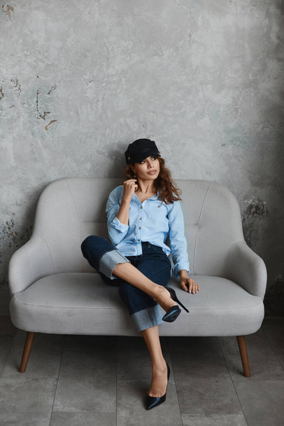 High fashioned model girl wearing high heel shoes, hat, blue shirt and roll-up jeans. Beautiful young woman in modish outfit sits on the couch and posing in the interior. Concept of street fashion. - Foto, imagen