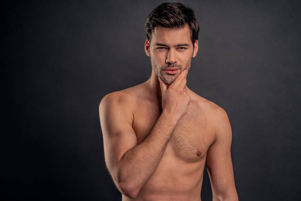 Handsome young bearded man isolated. Portrait of shirtless muscular man is standing on grey background and looking at camera. Holding his hand on chin. - 写真・画像