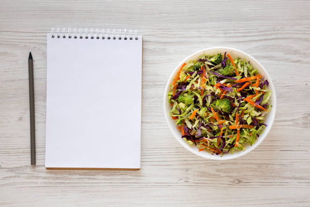 Homemade Raw Shredded Broccoli Slaw in a white bowl, blank notepad on a white wooden background, top view. Overhead, from above, flat lay. - Photo, Image