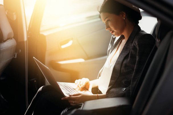 Businesswoman in a jacket sits in a car in the back seat and works at a laptop checking work mail - Фото, изображение