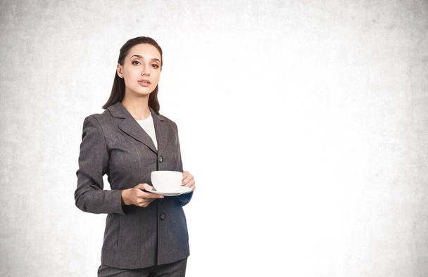 Portrait of beautiful young businesswoman in elegant suit holding white cup and saucer near concrete wall. Concept of coffee break and leadership. Mock up - Φωτογραφία, εικόνα