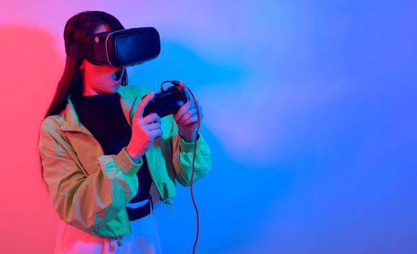 Ein schockiertes Mädchen in Neon mit VR-Brille hält ein Gamepad in der Hand und spielt Spiele. Virtuelle Welt, Technologie. - Foto, Bild
