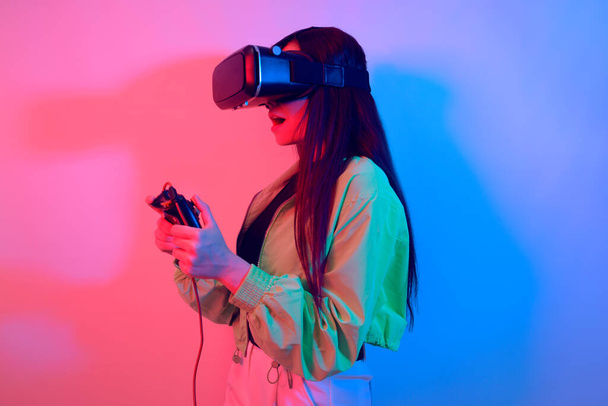 A shocked girl in neon with VR glasses holds a gamepad in her hands and plays games. Virtual world, technology. - Photo, Image