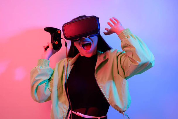 Laughing smiling surprise girl in neon with VR glasses holds a gamepad in her hands and plays games. Virtual world, technology. - 写真・画像