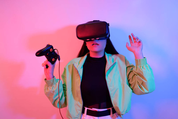 A frightened girl in neon with VR glasses holds a gamepad in her hands and plays games. Virtual world, technology. Emotions of fear, horror, horror games - Photo, Image