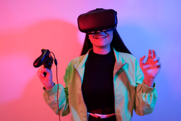 Lachend lachend verrassingsmeisje in neon met VR bril houdt een gamepad in haar handen en speelt spelletjes. Virtuele wereld, technologie. - Foto, afbeelding