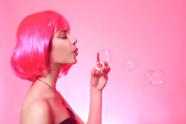 Chica con pelo rosa, cabello castaño. Modelo infla una burbuja de jabón, una mujer sobre un fondo rosa
. - Foto, Imagen