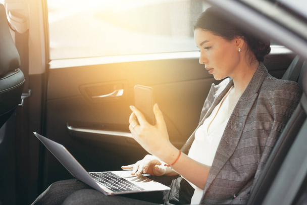 Mulher bonita em um terno de negócios senta-se no compartimento do passageiro e verifica os dados através de um smartphone e laptop
 - Foto, Imagem