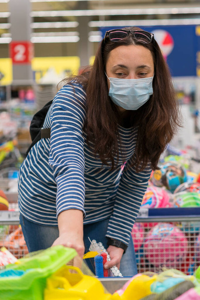 La donna in una maschera protettiva in un supermercato sceglie giocattoli da bambini. tonica. foto verticale. - Foto, immagini