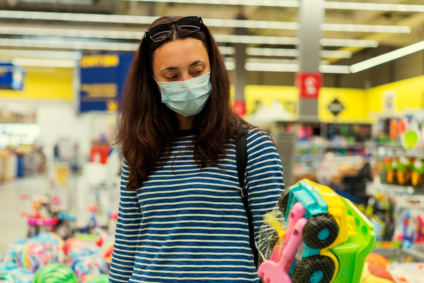 Frau mit Schutzmaske im Supermarkt wählt Kinderspielzeug. gemildert. - Foto, Bild