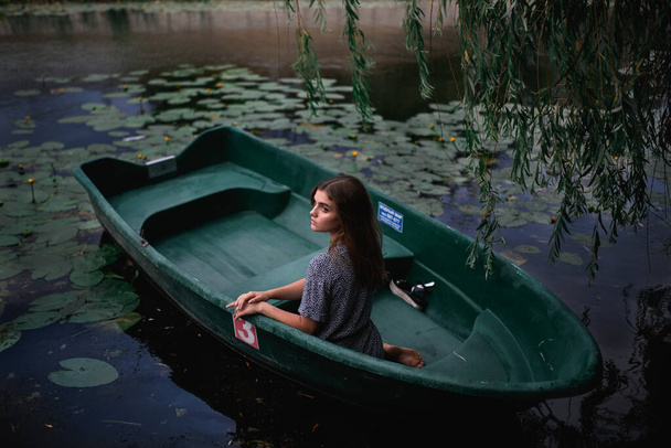 Een jong meisje zit in een oude boot tegen een achtergrond van waterlelies op een zomerdag - Foto, afbeelding