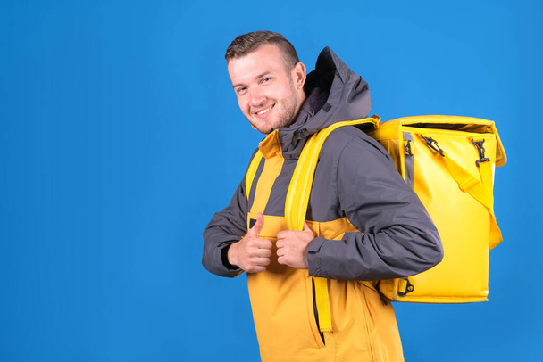 Young smiling caucasian food delivery guy in yellow uniform and refrigerator bag on his back waves finger up in approval or husky on blue background. Courier delivers groceries home. - Photo, Image