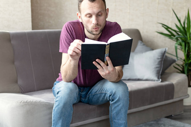 Nuori mies istuu sohvalla ja lukee kirjaa kiinnostuneena - Valokuva, kuva