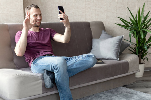 joven feliz hombre en una camiseta y jeans sentado en el sofá con un teléfono inteligente en la mano y hablando a través de videollamada
 - Foto, Imagen