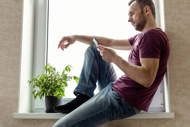 Tişört ve kot pantolonlu genç adam büyük bir pencerenin penceresinde oturur ve akıllı telefona bakar. - Fotoğraf, Görsel