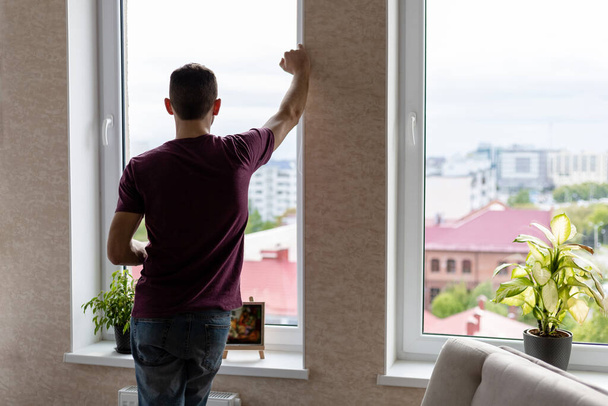 Młody człowiek w t-shircie i dżinsach stoi przy dużym oknie i patrzy na miasto - Zdjęcie, obraz