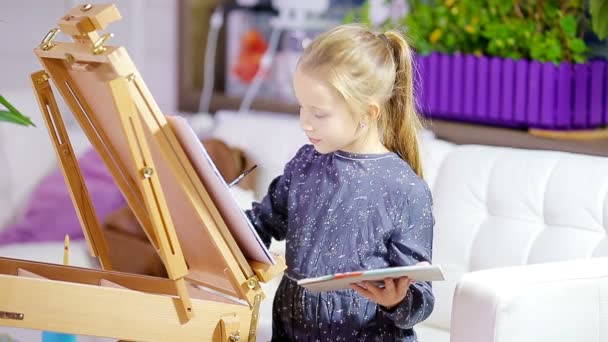 Urocza dziewczynka maluje obraz na sztalugach wewnątrz - Materiał filmowy, wideo