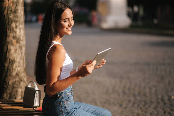 Beautiful girl use tablet outdoors. Female sitting on the bench alone. Woman watch video on tablet and smile. Space for text - Fotografie, Obrázek