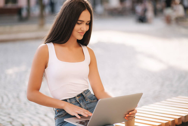 Attractive young woman use laptop outside on the bench in summer time. Woman working online outdoors. Alone. Beautiful brunette female. Student study - Photo, Image