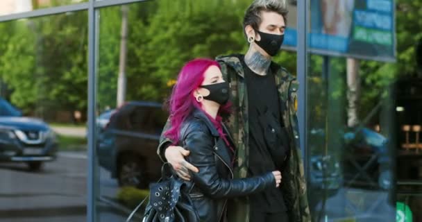 Vidám kaukázusi fiatal divatos pár hipszter sétál éhesen az utcán a kovid-19-es járvány idején. Gyönyörű rendkívüli barátnő rózsaszín haj és a barátja tetoválások sétáló maszkok - Felvétel, videó
