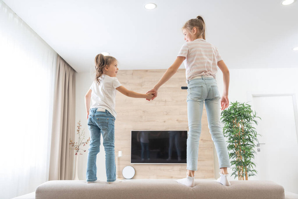 Two sisters jumping on couch in living room having fun - Photo, Image