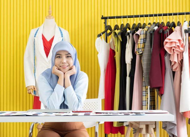 ビジネスのオーナーとしてヒジャーブを身に着けていた若いアジア系ムスリム女性デザイナーは、スタジオの作業台で喜んで笑っています. - 写真・画像