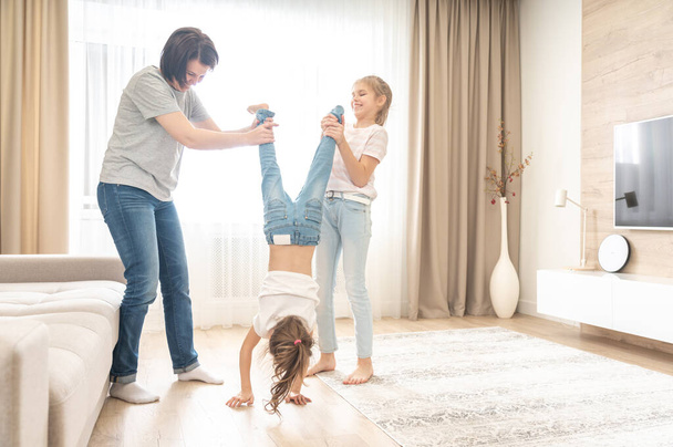 Щаслива мати і дві доньки розважаються у вітальні. мама тримає молодшу дочку догори ногами
 - Фото, зображення