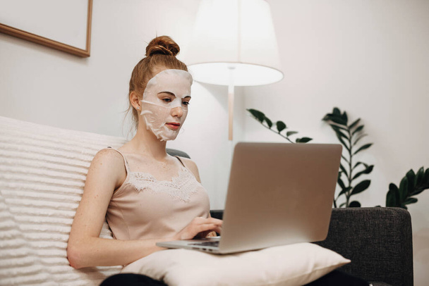 Fehér nő vörös hajjal, papírmaszkot visel az arcán, miközben fekszik az ágyban és laptopot használ. - Fotó, kép