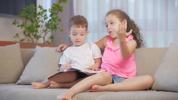 Cute children,sister with a younger brother, looking at a book magazine textbook dictionary tablet read fairy tales, kids are reading books sitting on the couch,preschool and school education. - Materiaali, video